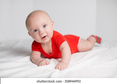 Portrait Of A Smiling 5 Months Baby Boy In A Red Onesie Lying Down On A White Blanket