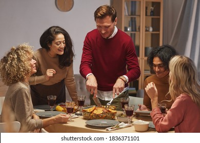 Portrait of smiling adult man cutting delicious roasted turkey while enjoying Thanksgiving dinner with friends and family, copy space - Shutterstock ID 1836371101