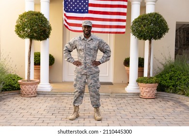 Portrait Of Smiling African American Army Man Standing With Hand On Hip Against Flag On House. Patriotism And Identity, Unaltered.