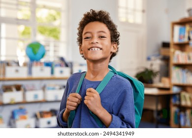 Portrait of smiling african american boy looking at camera while wearing green backpack. Black elementary schoolboy carrying backpack and standing in library at primary school.  - Powered by Shutterstock