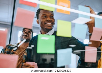 Portrait of a smiling african american businessman working on a new project and writing main topics while using colorful sticky notes on the glass wall. Shot trough a glass wall. Copy space. - Powered by Shutterstock