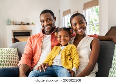 Portrait Of Smiling African American Family Cuddling On Sofa. Happy Mature Black Man And Woman Sitting On Sofa At Home With Daughter Looking At Camera. Happy Mother And Father With Little Girl.