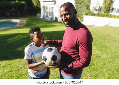 Portrait Of Smiling African American Father With Son Embracing And Holding Football In Sunny Garden. Family Spending Time At Home.