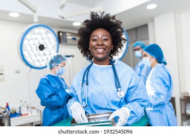 Portrait of smiling African American female surgeon in surgical uniform taking surgical instruments at operating room. Young woman doctor in hospital operation theater - Shutterstock ID 1932545096
