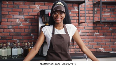 Portrait Of Smiling African American Female Barista Wearing Uniform At Workplace. Happy Woman Employee Standing Behind Cafe Counter Welcoming Visitors.