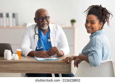 Portrait Of Smiling African American Female Patient Receiving Medical Consultation And Looking Back At Camera On Foreground, Mature Male Doctor Sitting At Desk In The Blurred Background