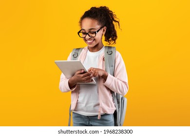 Portrait of smiling African American girl in glasses holding and using digital tablet, wearing backpack, browsing internet or doing homework standing isolated over yellow studio background - Powered by Shutterstock
