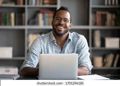 Portrait of smiling African American man in glasses sit at desk in office working on laptop, happy biracial male worker look at camera posing, busy using modern computer gadget at workplace - Shutterstock ID 1701296185