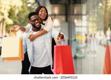 Portrait of smiling African American man giving piggyback ride to his happy woman who holding shopper bags, couple looking and pointing at shop window, walking near shopping center