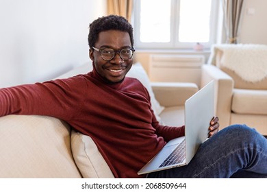 Portrait of smiling African American man in glasses sit at sofa in office working on laptop, happy biracial male worker look at camera posing, busy using modern computer gadget at workplace