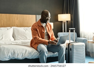 Portrait of smiling African American man using laptop sitting on bed in hotel room and researching guest services copy space - Powered by Shutterstock