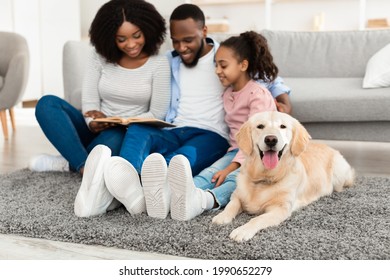 Portrait Of Smiling African American Mum, Dad And Daughter Reading Paper Book Or Looking At Photo Album Together Sitting On The Floor Carpet In The Blurred Background, Selective Focus On Happy Dog