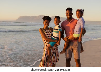 Portrait Of Smiling African American Parents Carrying Son And Daughter While Standing Against Sea. Nature, Unaltered, Beach, Childhood, Family, Togetherness, Lifestyle, Enjoyment And Holiday.