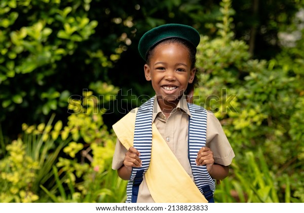 Portrait of smiling african\
american scout girl in uniform with backpack against plants.\
unaltered, girl scout, childhood, courage, leadership and\
scouting.