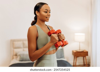 Portrait of smiling African American sportswoman in sportwear working out with hand weights in bedroom at home. Sports, health care concept - Powered by Shutterstock