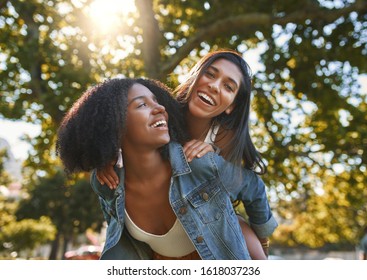 Portrait Of A Smiling African American Young Woman Carrying Her Best Friend On Her Back Giving Piggyback Ride In Park On A Sunny Day - Friends Laughing And Having Fun Outdoors