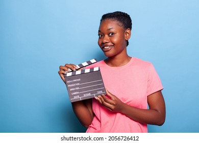 Portrait of smiling african american young woman holding movie production blackboard standing in studio witg blue background. Teenager giving audition for movie roll. Cinematographic concept