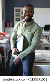 Portrait of smiling african american young male teacher with file sitting at desk in classroom. unaltered, education, teaching, occupation and school concept.
