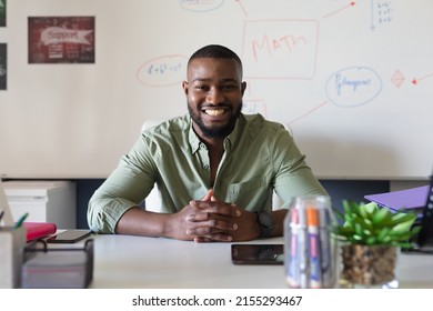 Portrait Of Smiling African American Young Male Teacher Sitting At Desk Against Whiteboard In Class. Unaltered, Education, Teaching, Occupation And School Concept.