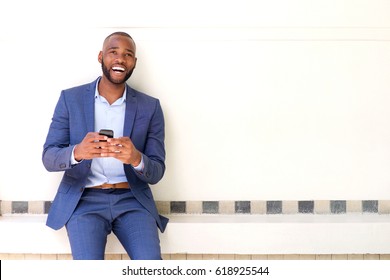 Portrait Of Smiling African Business Man Sitting Against Wall With Mobile Phone