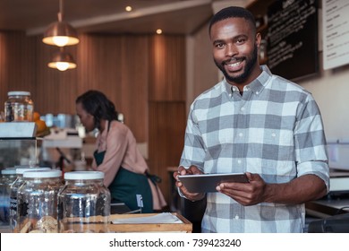 Portrait of a smiling African entrepreneur standing behind his cafe counter using a digital tablet with a colleague working in the background
