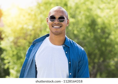 Portrait Of Smiling African Man Looking At Camera With Sunglasses. Happy Guy Enjoying The Summer Outdoor. Cheerful Young Man Wearing White Tshirt And Blue Shirt At Park. 