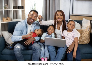 Portrait Of Smiling African Parents With Their Three Kids Holding Keys From New House. Happy Family Sitting On Couch With Modern Laptop Among Many Boxes.