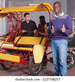 Portrait of smiling African-American pedicab driver standing near rickshaw cycle