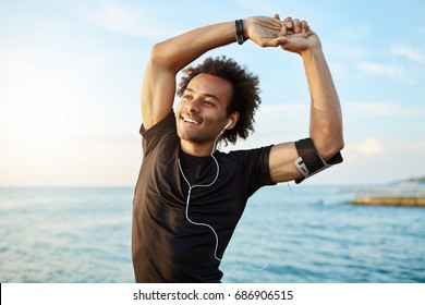 Portrait of a smiling afro-american sports man stretching his muscular arms before workout by the sea, using music app on his smartphone. Dark-skinned athlete warming up before running. - Powered by Shutterstock