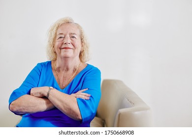 Portrait of smiling aged woman in blue dress crossing arms and looking at camera when sitting in chair - Shutterstock ID 1750668908