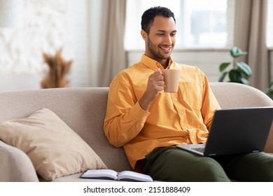 Portrait of smiling Arabic bearded man using laptop, holding cup and enjoying drinking morning coffee. Adult male sitting on comfy couch in living room, watching video, taking break, free copy space - Powered by Shutterstock