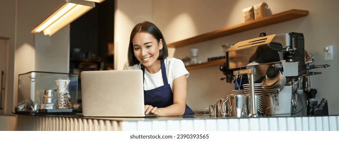 Portrait of smiling asian barista, cafe owner entrepreneur, working on laptop, processing orders on computer, standing behind counter. - Powered by Shutterstock