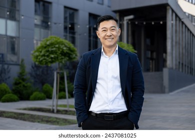 Portrait of a smiling Asian businessman in a suit standing confidently outside a contemporary office complex on a sunny day. - Powered by Shutterstock