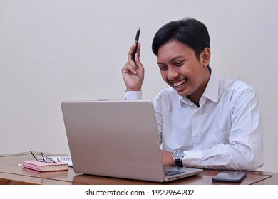 Portrait Of Smiling Asian Businessman In White Shirt Laughing While Looking At Laptop Screen And Holding A Pen. Employee Working In The Office. Isolated Image Over Gray Background