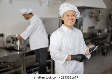 Portrait of smiling asian chef in uniform standing with printed order in professional kitchen with latin guy cooking on background - Powered by Shutterstock