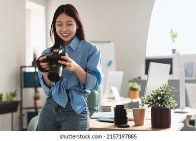 Portrait Of Smiling Asian Female Photographer Holding Camera Working With Photos, Reviewing Looking Through Taken Pictures At Office, Sitting Leaning On Table Desk. Art And Creative Profession Concept - Powered by Shutterstock