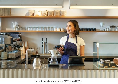 Portrait of smiling asian girl barista, standing near coffee brewing kit, making filter in cafe, holding POS terminal, processing clients order. - Powered by Shutterstock