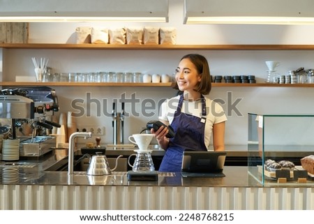 Portrait of smiling asian girl barista, standing near coffee brewing kit, making filter in cafe, holding POS terminal, processing clients order.