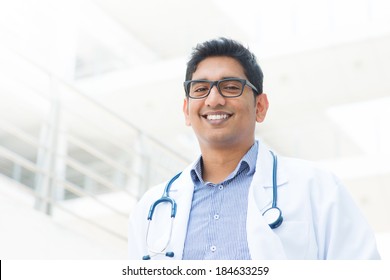 Portrait Of A Smiling Asian Indian Male Medical Doctor In Uniform, Hospital Building At Background.