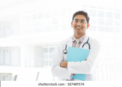 Portrait Of A Smiling Asian Indian Male Medical Doctor Standing Inside Hospital, Holding File Folder.