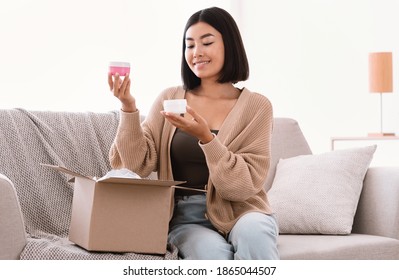 Portrait Of Smiling Asian Lady Received Package, Unpacking Cardboard Beauty Box With Cosmetics Product, Holding Skincare Cream In Both Hands Sitting On The Couch. Woman Satisfied With Online Shopping