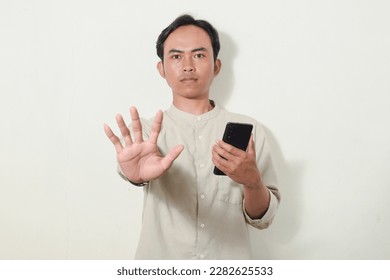 portrait of a smiling Asian man holding a smartphone and showing palms. portrait of indonesian man in gray shirt looking at camera on isolated white background. gestures stop, refuse, don't, prohibit - Powered by Shutterstock