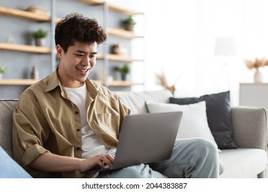 Portrait Of Smiling Asian Man Sitting On The Couch Working On Pc Laptop Indoors In Living Room. Happy Male Using Computer For Rest, Study Or Education, Typing On Keyboard, Surfing Internet