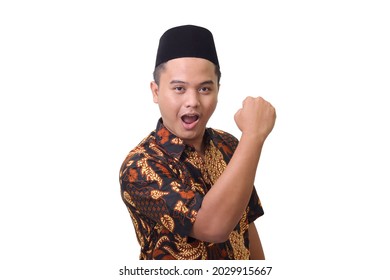 Portrait of smiling Asian man wearing batik shirt and songkok raising his fist, celebrating success. Isolated image on white background