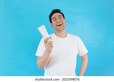 Portrait Smiling Asian Man In White T-shirt Holding Facial Cleanser Face Wash Soap Over Blue Background With Copy Space.