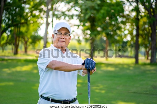 Portrait of Smiling Asian senior man golfer holding\
golf club standing on golf course in summer sunny day. Healthy\
elderly male enjoy outdoor lifestyle activity sport golfing at golf\
country club.