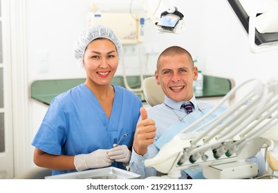 Portrait Of A Smiling Asian Woman Dentist With A Satisfied Young Man Patient Sitting In A Dental Chair In The Clinic Office