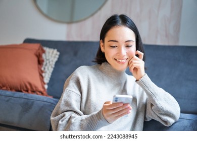 Portrait of smiling asian woman looking intrigued at smartphone screen, interested with smth on mobile phone, sitting coy on floor in living room.