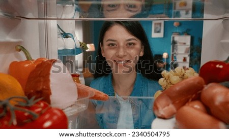 Portrait of a smiling Asian woman at the open refrigerator. A woman is satisfied with the abundance of food in the refrigerator. View from inside the refrigerator.