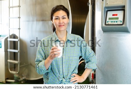 Portrait of a smiling asian woman standing in a dairy farm workshop with a glass of milk, showing off production...Close-up portrait